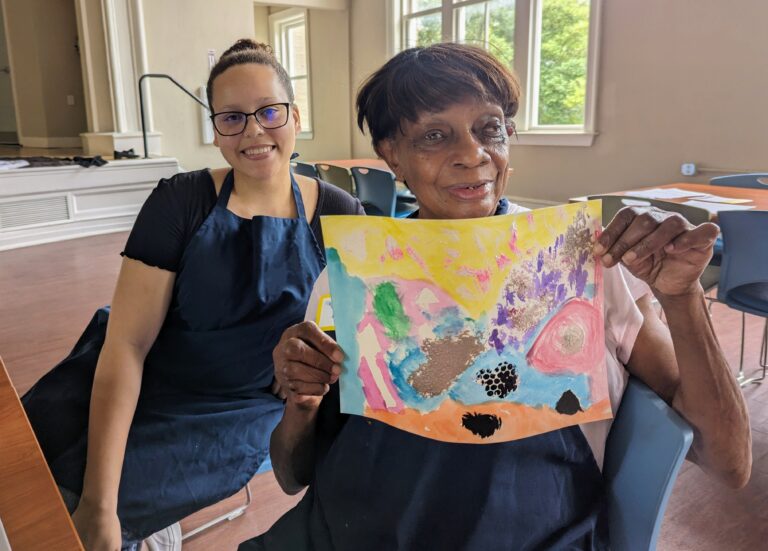 Older adult participant shows her painting with a volunteer.