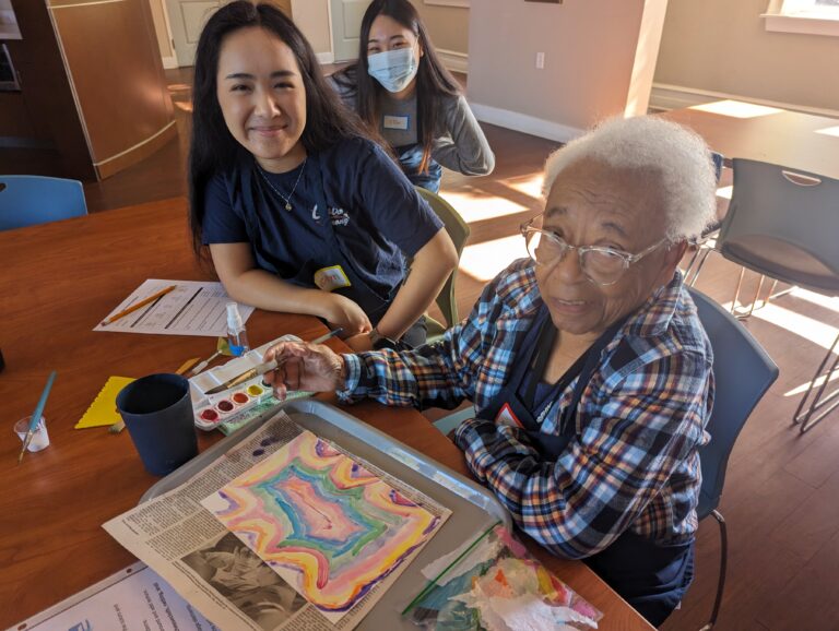 Two volunteers assist older adult participant in painting abstract art.