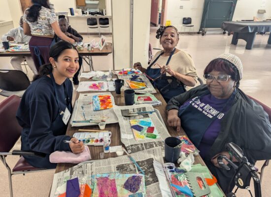 Two older participants and a volunteer do a newspaper painting activity.