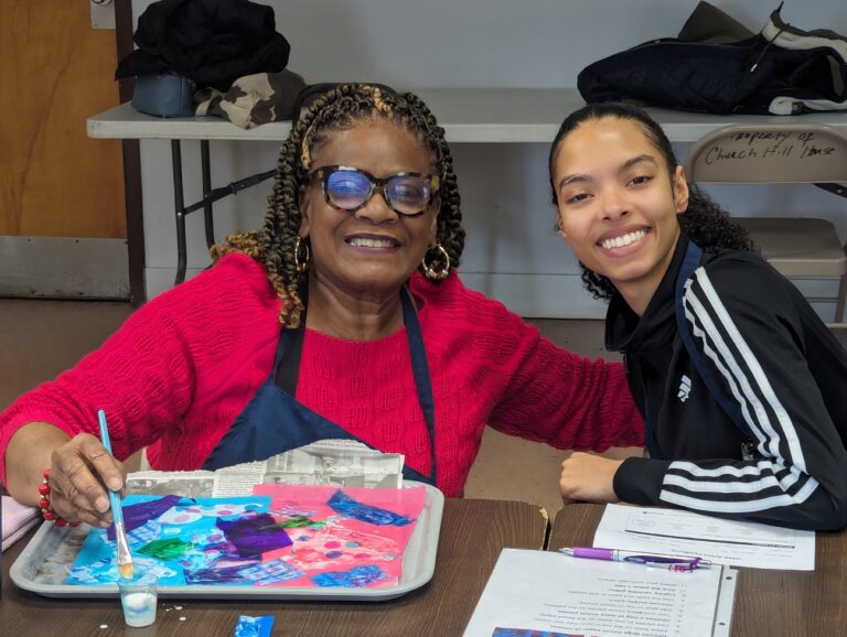 Older lady participant poses with volunteer as she paints on newspaper.