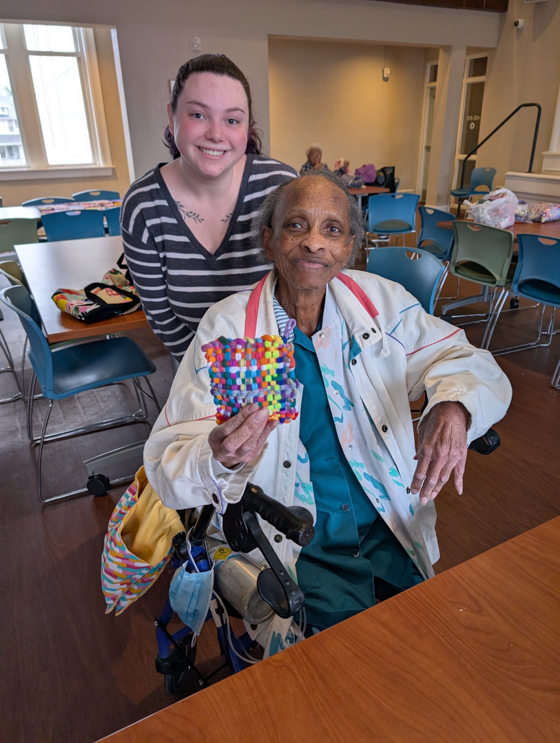 Older adult participant sitting on rollator shows off custom crochet square with volunteer.